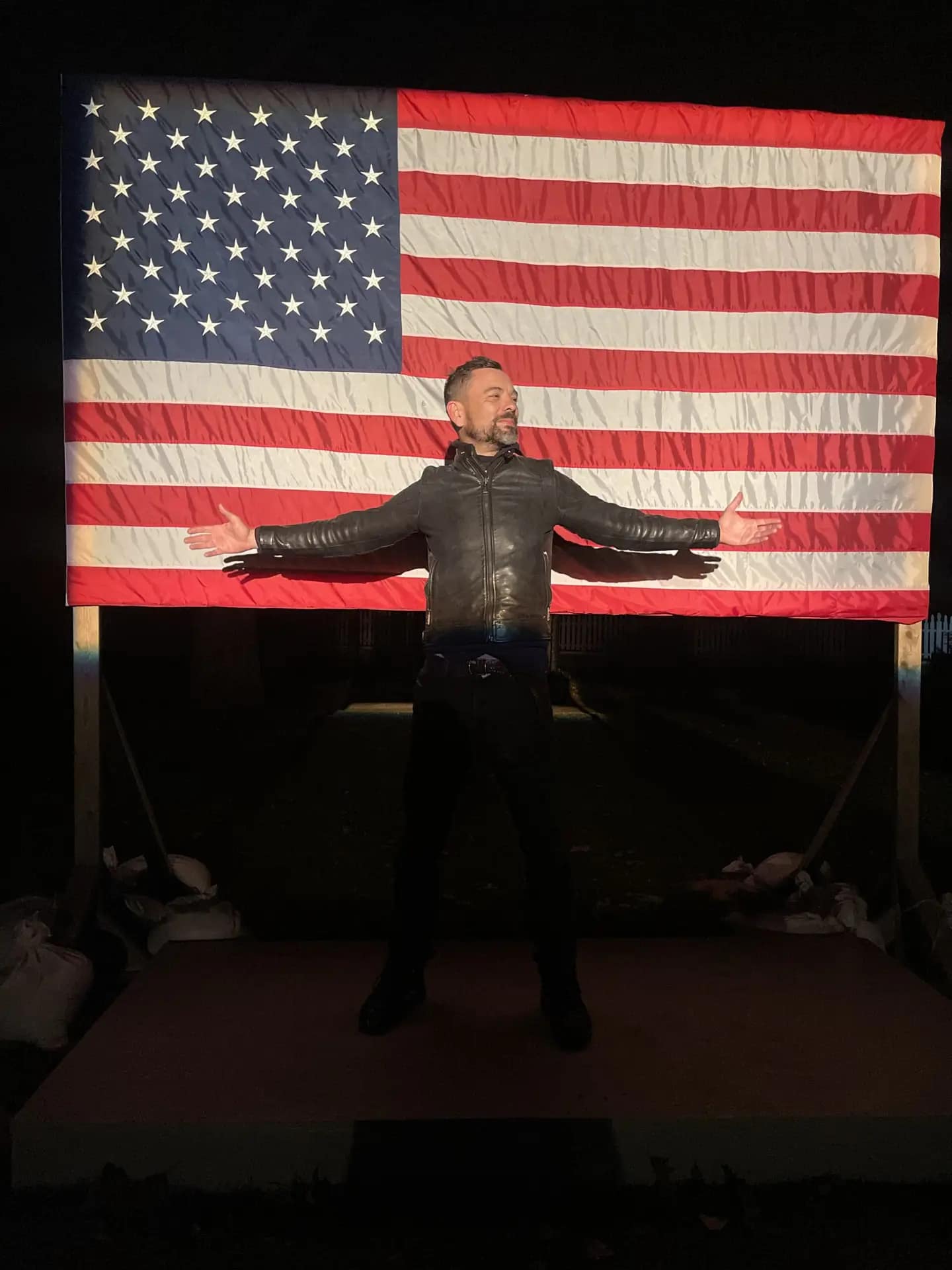 Man standing with arms outstretched in front of a large American flag.