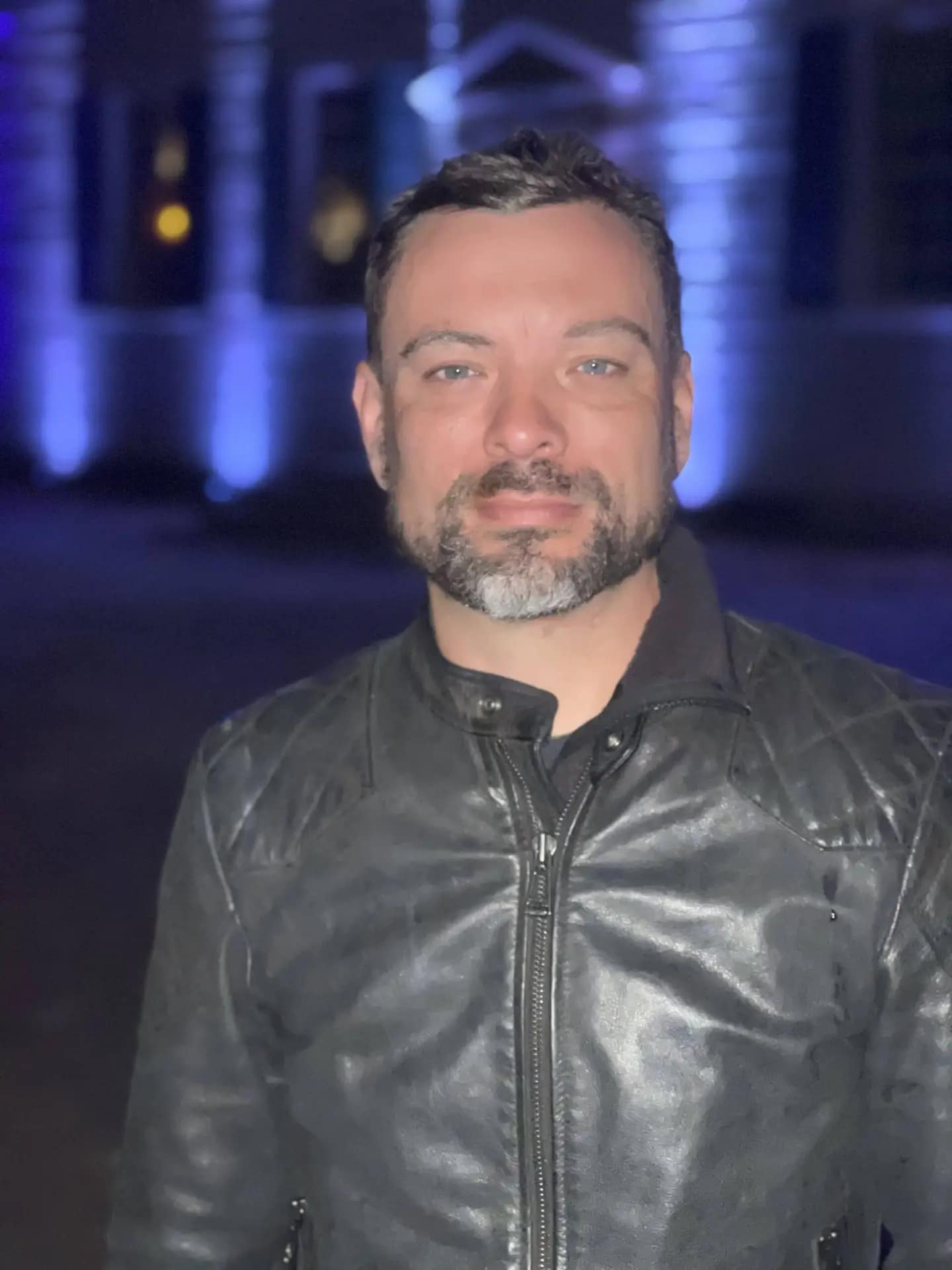 Man in a black leather jacket smiling in a night setting.