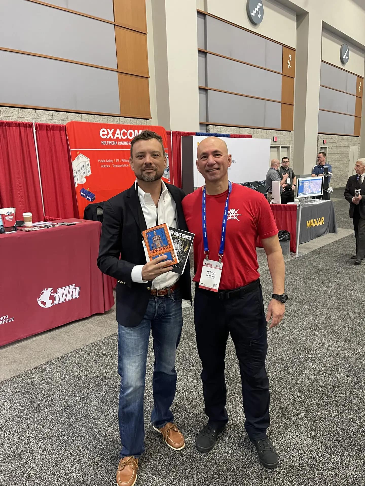Two men posing at a conference booth, one holding books.