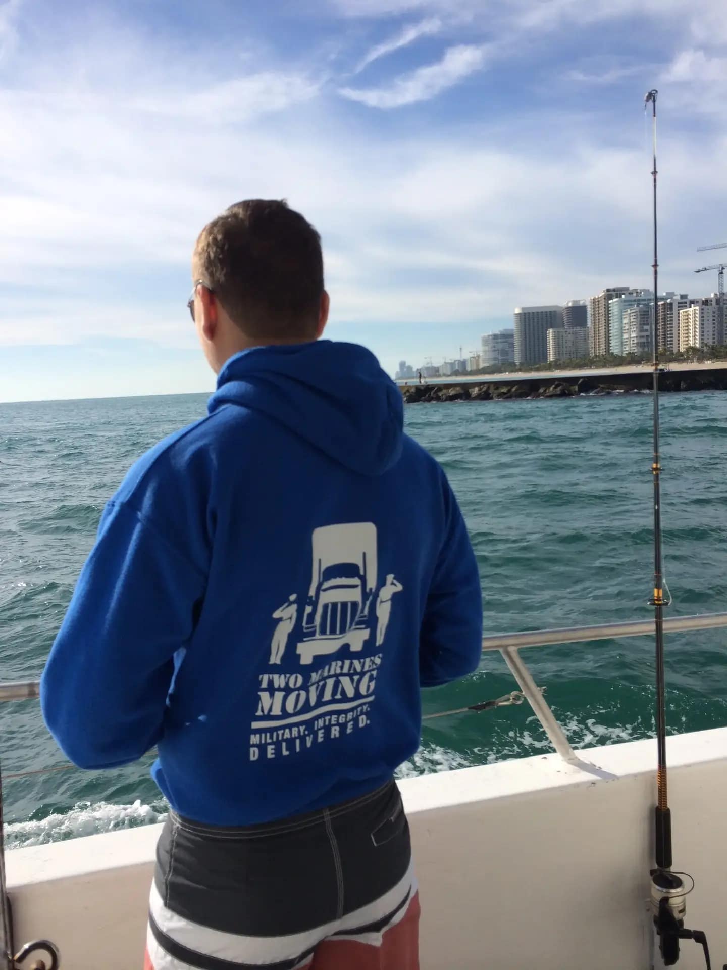 Person fishing on a boat near a city coastline with cloudy sky.