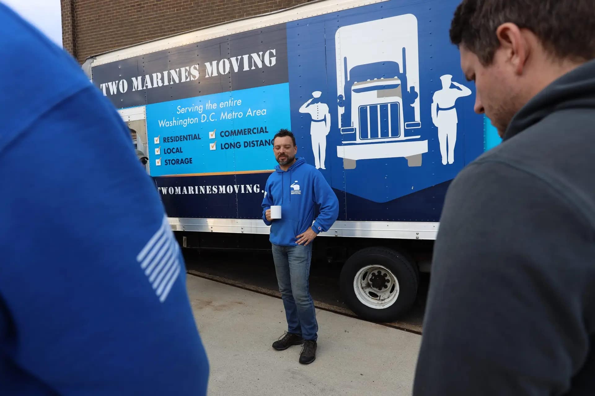 A man speaks to a group near a moving truck outside a building.