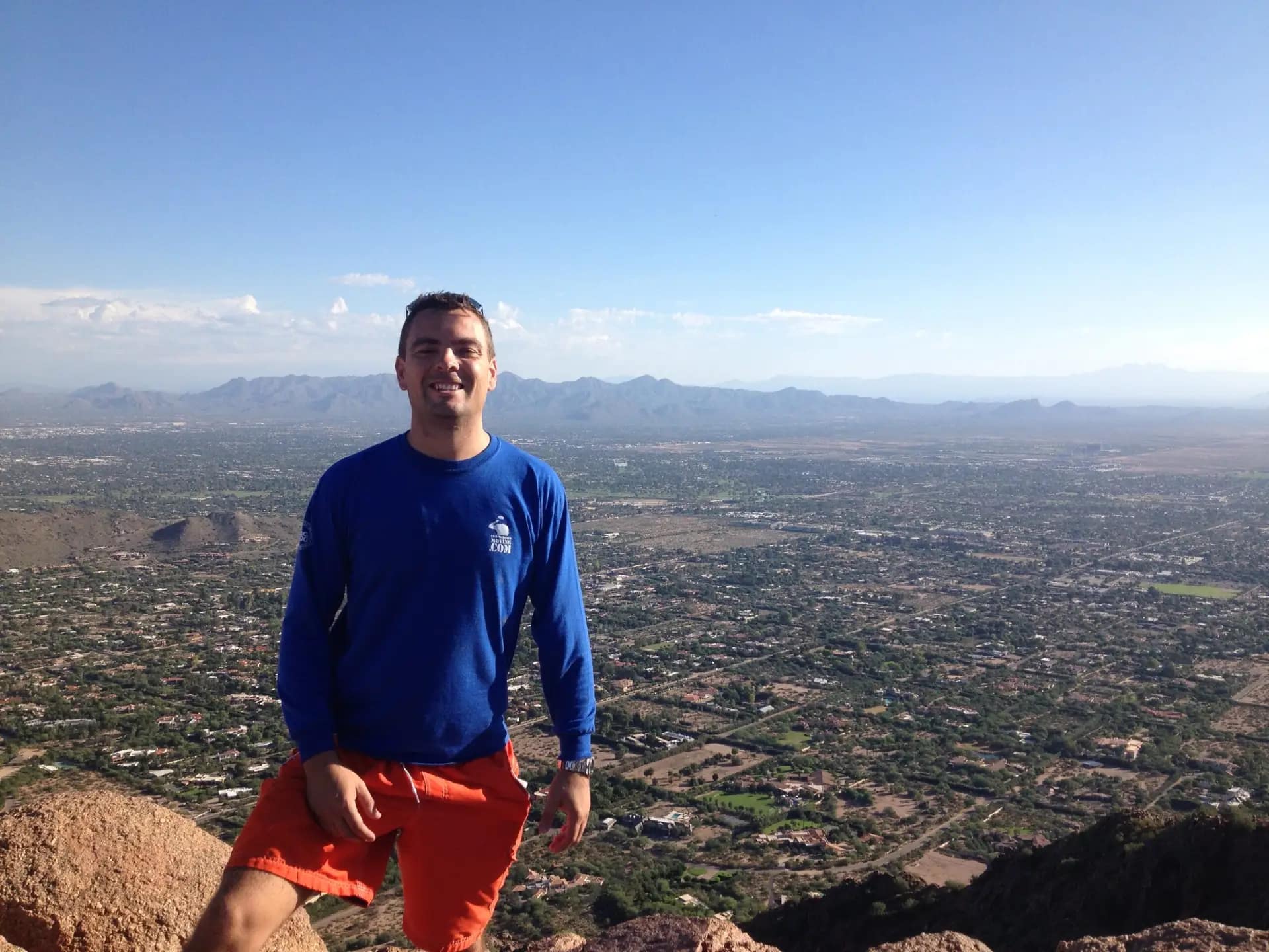 Man in blue sweater and red shorts stands on a mountain with a cityscape below.