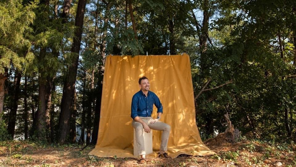 Man sitting on a drum in front of a mustard-colored backdrop in the woods.